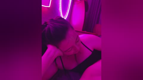 A video of a smiling woman with medium-length dark hair, wearing black lingerie, lying on a bed with pink and purple neon lights in the background.