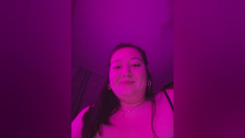 Video of a plus-sized woman with long dark hair, wearing a black top, under purple lighting, smiling slightly, with a soft focus.
