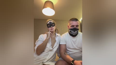 Video of a couple in a dimly-lit room wearing black face masks; woman with blonde hair, man with short hair.