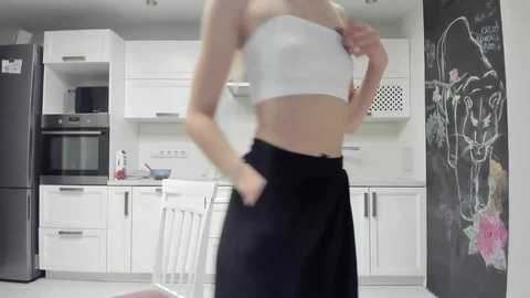 Media: A video of a woman in a modern kitchen, wearing a white crop top and black skirt, standing next to a chalkboard wall and white chairs.