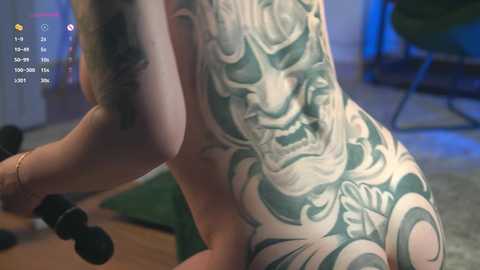 Video of a nude woman with a large, detailed green and black tattoo on her back, exercising in a dimly lit room with weights on a green mat.