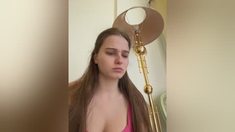 Video of a young woman with long, straight brown hair, wearing a pink tank top, standing in a room with beige walls. She has a neutral expression. A gold floor lamp is partially visible behind her.