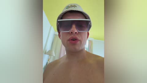 Video of a shirtless young man with fair skin, wearing large dark sunglasses and a white baseball cap, indoors with yellow walls and a lamp in the background.