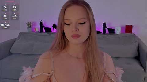 Video of a fair-skinned, blonde woman with long hair, wearing a light pink, off-shoulder top, sitting on a gray sofa in a dimly lit room with purple ambient lighting and black candle holders.