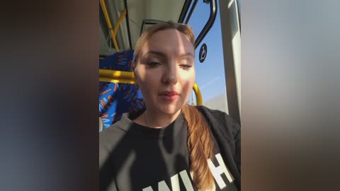Video of a young woman with fair skin and light brown hair, wearing a black t-shirt, seated on a bus, with sunlight casting shadows.