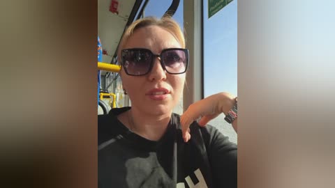 Video of a blonde woman with sunglasses, wearing a black shirt, sitting in a bus, looking out the window.