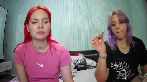 Media: Video of two young women with vivid red and purple hair, sitting on a bed with a messy background. One wears a pink Nike T-shirt, the other a black shirt with a leaf design.