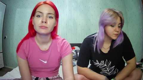 Media: Video of two young women with vibrant red and lavender hair, sitting on a bed in a room with green striped wallpaper. One wears a pink Nike shirt, the other a black graphic tee.