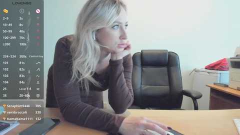A video of a blonde woman in a brown sweater, leaning on her hand, sitting at a desk in a minimalist office.
