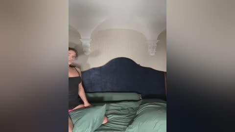 Media: Video of a woman with light skin and short dark hair, wearing a black tank top, standing on a green bed in a dimly lit room with an arched ceiling and a dark blue headboard.