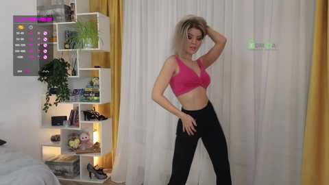 Media: Video of a blonde woman in a pink bra and black leggings, posing confidently in a modern, minimalist bedroom with white walls and yellow curtains.