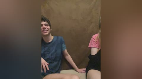 Video of a young man with short brown hair, wearing a dark blue t-shirt and shorts, sitting on a bench, grinning. A partially visible woman in a pink t-shirt and black shorts sits beside him.