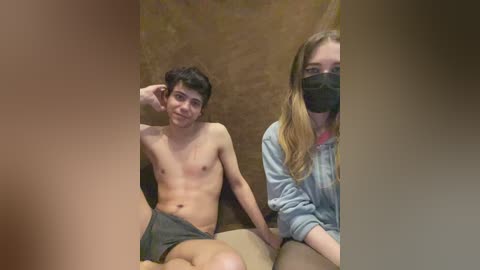 Video of two young people in casual attire: a shirtless teen with short dark hair in gray shorts and a masked young woman with long blonde hair, sitting on a beige sofa.