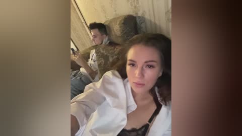 Media: Video of a young woman with fair skin and long brown hair, wearing a white shirt and black lace bra, sitting on a couch in a dimly lit room, next to a man with dark hair, holding a phone.