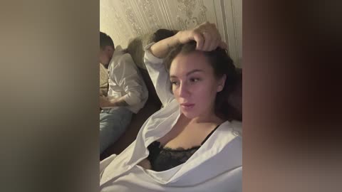 Media: A video shows a woman with brown hair, wearing a white shirt and black lace bra, sitting on a couch, with a man in the background, both looking at a phone.