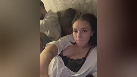 Media: A video of a young woman with medium skin tone and dark hair, wearing a white robe over a black lace bra, lying on a bed with a gray wall and patterned wallpaper in the background.