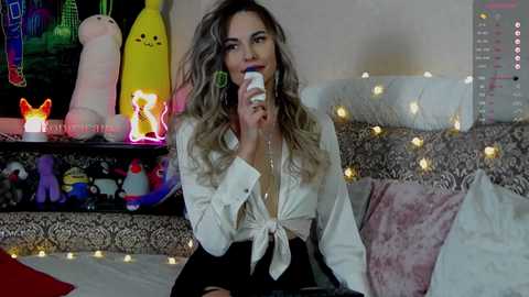Video of a woman with wavy, light brown hair, wearing a white blouse tied at the waist, and black skirt, holding a phone, surrounded by colorful plush toys and fairy lights in a cozy bedroom.