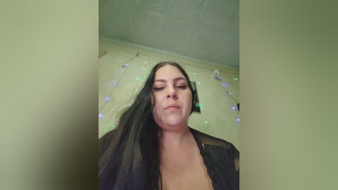 Video of a woman with long black hair, wearing a black robe, standing in a room with a textured ceiling and fairy lights.