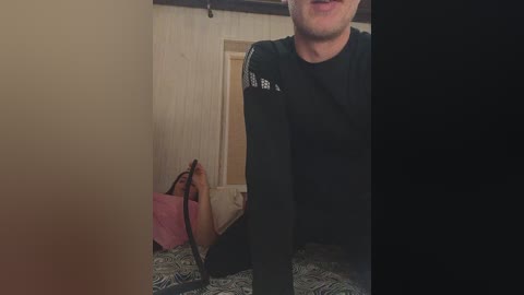 Media: Video of a man with a black long-sleeve shirt, holding a pink handbag, standing in a dimly lit room with a white door and patterned carpet.