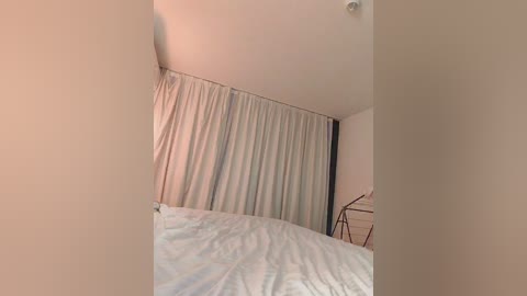 Media: Video of a minimalist bedroom with beige walls, a white bed with a light blue comforter, and a floor-to-ceiling white curtain.