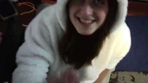 Media: Video of a young woman with long brown hair, wearing a white hoodie, smiling from above, against a blurred indoor background with a blue mat and orange wires.