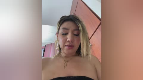Video of a young woman with shoulder-length blonde hair, wearing a black strapless top, standing in a room with pink walls and a wooden cabinet, looking downward.