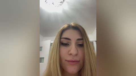 A video of a young, fair-skinned woman with straight, blonde hair, wearing makeup, looking down, with a dimly lit ceiling and white walls in the background.