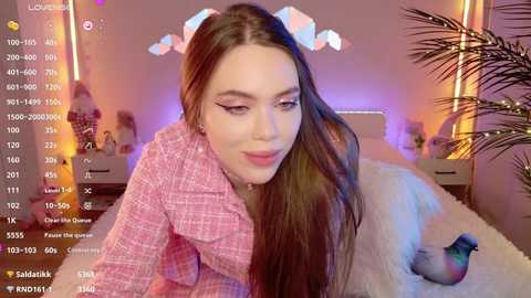 Media: Video of a young woman with long brown hair, wearing a pink plaid shirt, sitting on a white bed with a fluffy pillow, in a cozy room with pink and purple lighting and a palm tree.