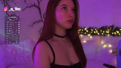 Video of a young woman with long red hair, wearing a black choker necklace and a black lace top, seated in a dimly lit room with purple lighting and Christmas decorations.