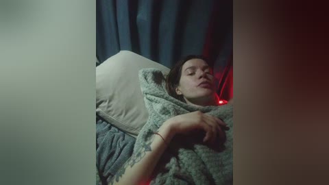 Video of a young woman with fair skin, brown hair, and a tattoo on her left forearm, lying in bed, covered in a gray blanket, against dark blue curtains.