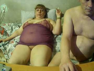 Video of an overweight, middle-aged woman in a purple nightgown, lying on a bed with a patterned blanket, and a thin, nude man beside her.