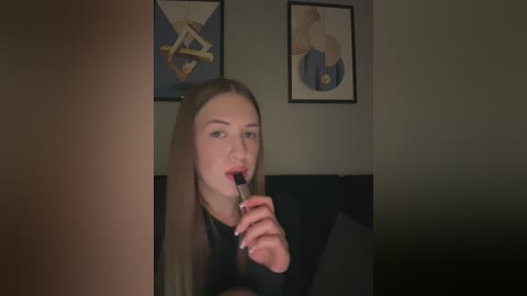 Media: A video of a young Caucasian woman with straight, long blonde hair, wearing a black top, licking a lollipop in a dimly lit room. Behind her are two framed, abstract art pieces on a grey wall.