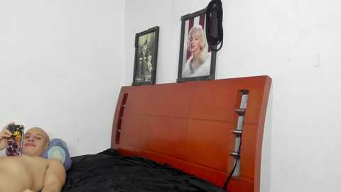 Video of a nude, bald man lying on a bed with a red wooden headboard, surrounded by black sheets and framed Marilyn Monroe posters on white walls.