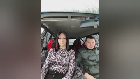Media: Video of a woman with straight black hair, wearing a leopard-print top, sitting next to a man in a green puffer jacket, inside a car with red seats and a grey interior.
