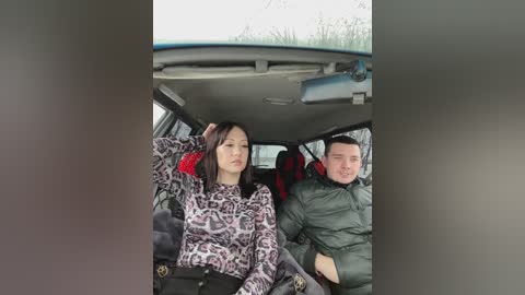 Media: Video of two people in a car: a woman with black hair, wearing a leopard print top and black pants, and a man in a green jacket, both looking tired and bored.