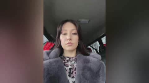 Media: A video of an Asian woman with long black hair, wearing a leopard print top, a grey fur coat, and sitting in a car. The background shows a cluttered interior with hanging clothes.