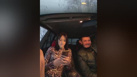 Media: Video of a woman with long brown hair in a leopard print dress, sitting next to a man with short brown hair in a black jacket, both smiling and taking selfies in a car during daylight.