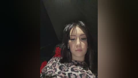 Media: Video of a young woman with long dark hair, wearing a leopard print top, sitting on a chair, with a red patterned cushion behind her, in a dimly lit room.