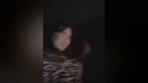 Media: A blurry video of a woman with dark hair, wearing a patterned jacket, in a dimly lit, dark room. Her face is partially obscured by shadows.