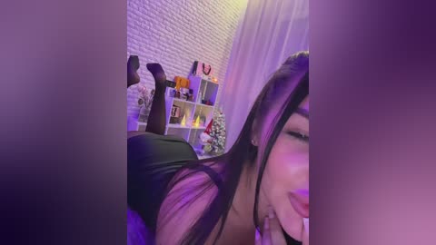 Media: Video of a woman lying on her stomach, wearing black stockings and high heels, in a dimly lit room with purple lighting, a white brick wall, and a shelf with various items.