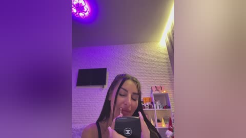 Media: A video of a young woman with long black hair, fair skin, and a slender physique, taking a selfie in a dimly lit room with a brick-patterned wall.