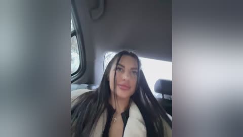 Media: Video of a woman with long, straight black hair, wearing a white jacket and black top, seated in a car with the window down, revealing a blurred view of the outside.