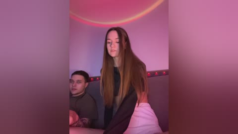 Media: Video of a young Caucasian woman with long brown hair, wearing a black top and white pants, standing in a dimly lit room with pink lighting and a man seated behind her.