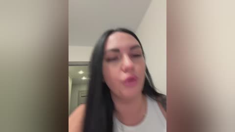 A blurry video of a woman with long black hair, wearing a white tank top, making a kissing face in a dimly lit hallway with white walls.