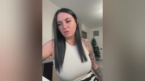 Video of a woman with straight black hair, fair skin, and a sleeve tattoo, wearing a white tank top, making a face in a modern, minimalist room with a Christmas tree in the background.
