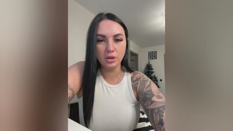 Video of a young woman with long black hair, wearing a white tank top, showcasing extensive tattoos on her right arm. She stands indoors with a Christmas tree and framed art in the background.