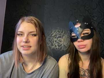 Video of two young women with long, straight brown hair. One wears a gray shirt, the other a black mask with a blue eye design. Background features a dark, textured wall with a circular white design.