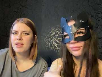Media: Video of two young women with light skin, one with straight, shoulder-length auburn hair, wearing a gray top, and the other with long, dark brown hair, wearing a black, decorative masquerade mask. Background features a dark, ornate wallpaper.