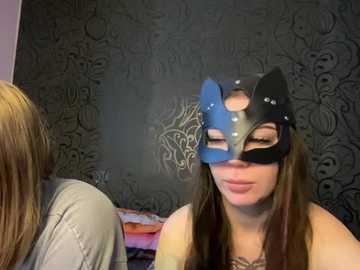 Media: A video shows a young woman with long brown hair, wearing a black cat mask with blue ears, standing in front of a dark, patterned wall. Another person's blonde hair is visible in the background.