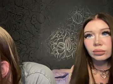 Video of a young woman with long brown hair, wearing a brown coat and heart necklace, standing next to a person with long blonde hair, in a room with dark, ornate wallpaper.
